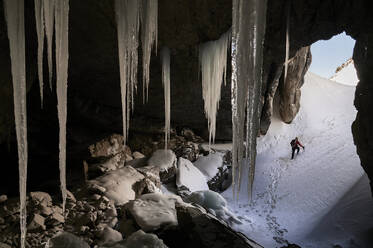 Back view of anonymous tourist with backpack and in warm clothes walking on snowy ground behind icicles while spending time alone at winter day in Garganta de Borau, Spain - ADSF44785