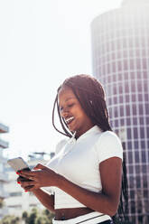 Positive African American female in casual clothes and with Afro braids smiling and browsing mobile phone while standing on city street on sunny day - ADSF44788