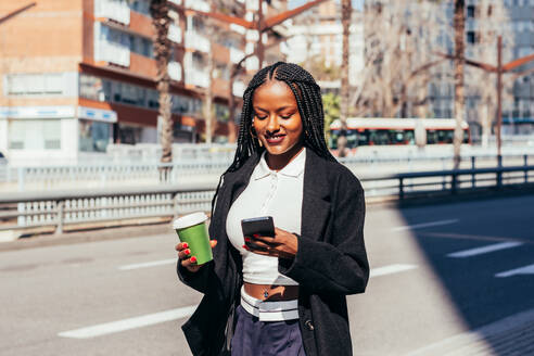 Positive African American female with braids smiling and looking at phone screen while standing with disposable cup of coffee on city street - ADSF44789