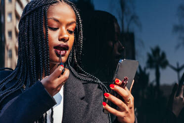 African American female with Afro braids standing on street and applying lipstick while looking at smartphone screen - ADSF44790