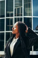 Young African American female with Afro braids and in black jacket standing on city street with hand on head and closed eyes - ADSF44795