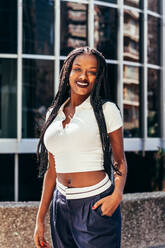 Happy young African American female with long Afro braids looking at camera while standing near modern building on city street - ADSF44797