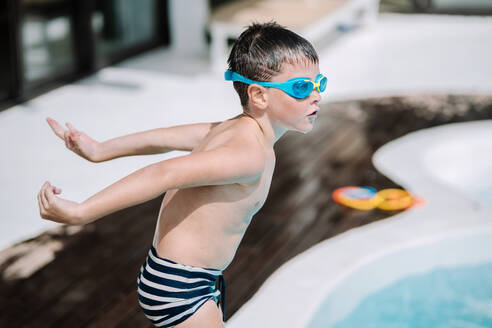 Kid in swimwear and diving glasses about to jump into swimming pool while enjoying summer vacation during holidays - ADSF44804