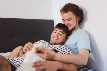 Happy young gay couple in domestic clothes lying on comfortable bed with dog and cuddling while smiling brightly and resting together during weekend at home - ADSF44812