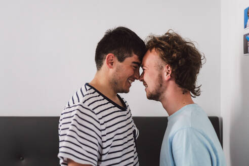 Side view of cheerful young gay couple in casual t-shirts smiling and looking at each other while standing near white wall at home - ADSF44814