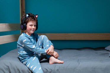 Smiling cute child with wireless kids headphones sitting on bed in nightdress and looking at camera while passing time and listening to music in bedroom - ADSF44817