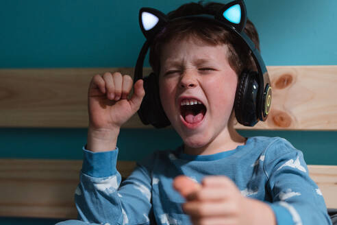 Happy kid in nightdress yelling with excitement with eyes and fists closed while listening to music in wireless headphones and leaning on wooden planks against blue background - ADSF44819