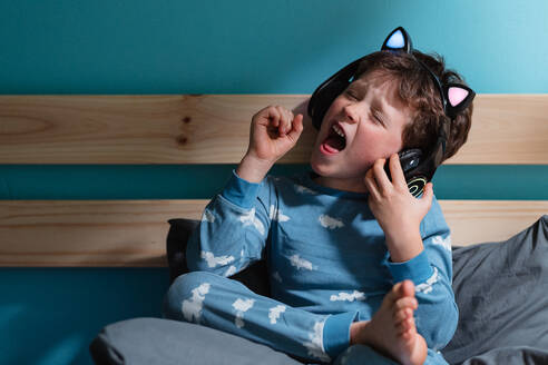 Happy kid in nightdress yelling with excitement with eyes and fists closed while listening to music in wireless headphones and leaning on wooden planks against blue background - ADSF44820