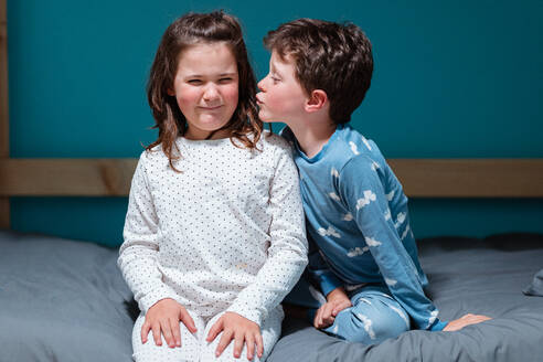 Cute siblings in pajamas sitting on bed while brother kid making kissing fun of sister child with eyes closed while enjoying free time before bedtime in bedroom - ADSF44821