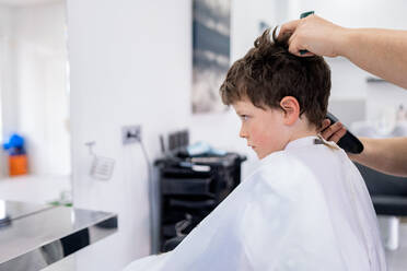 Unrecognizable hairdresser using comb and machine to trim hair of boy sitting in white cape in salon with blurred furniture - ADSF44825