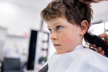 Unrecognizable hairdresser using comb and machine to trim hair of boy sitting in white cape in salon with blurred background - ADSF44826