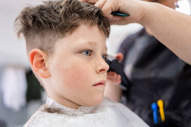 Unrecognizable hairdresser using comb and machine to trim hair of boy sitting in white cape in salon with blurred background - ADSF44827