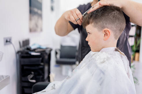 Side view of child sitting and wearing cape while anonymous hairdresser with comb and scissors doing hairdo in blurred salon - ADSF44829