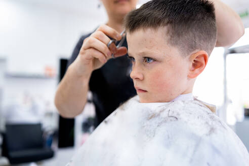 Side view of child sitting and wearing cape while anonymous hairdresser with comb and scissors doing hairdo in blurred salon - ADSF44830