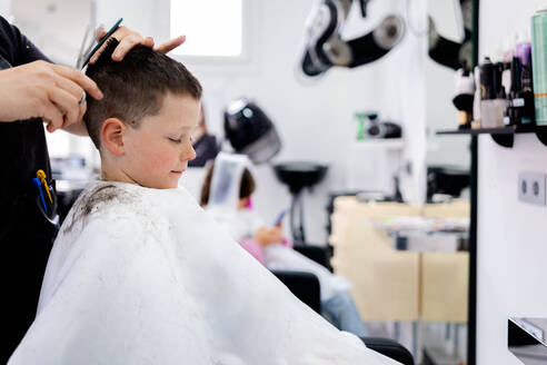 Side view of child sitting with eyes closed wearing cape while anonymous hairdresser with comb and scissors doing hairdo in blurred salon with clients - ADSF44831