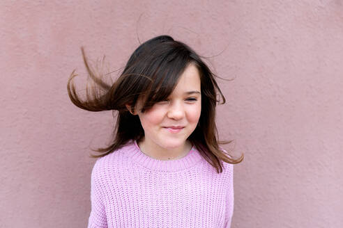Adorable cute child in sweater smiling and looking away with flying dark hair while standing against dark pink textured background - ADSF44833