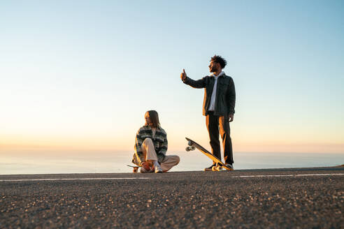 Full body of diverse happy young man and woman with skateboards and in casual clothes showing thumb up gesture to catch ride while standing in mountains against sea during sunset - ADSF44840