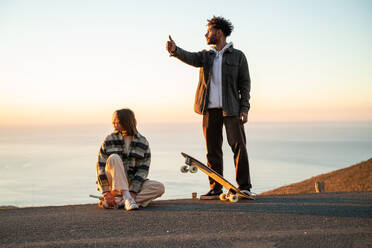 Full body of diverse happy young man and woman with skateboards and in casual clothes showing thumb up gesture to catch ride while standing in mountains against sea during sunset - ADSF44841