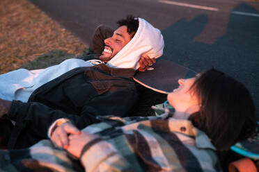 Young multiethnic couple in casual clothes and with skateboards lying on grass and laughing happily with closed eyes while relaxing together in nature - ADSF44842