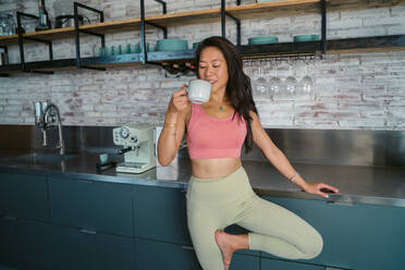 Young Asian smiling female athlete drinking cup of water while standing on tree pose near bar counter during break in fitness studio - ADSF44846