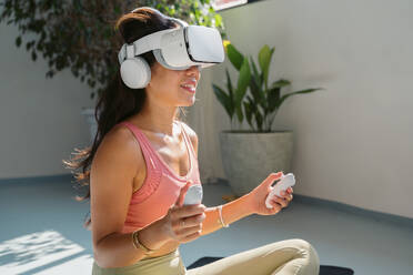 Young asian woman in sportswear sitting on mat and using virtual reality headset while doing fitness exercise during workout - ADSF44847