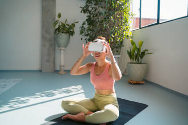 Full body of young barefoot female in activewear and virtual reality goggles sitting on yoga mat with crossed legs in light studio - ADSF44848
