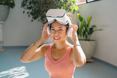 Cheerful asian female in activewear and virtual reality goggles smiling and looking at camera in light studio - ADSF44849