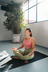 Full body of peaceful young Asian barefoot female in activewear sitting on yoga mat in lotus pose with Gyan mudra hands while meditating in modern light studio - ADSF44854