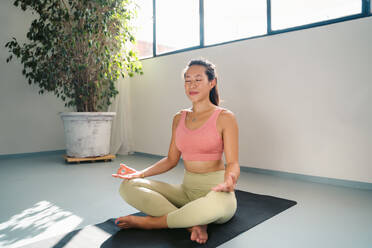 Full body of peaceful young Asian barefoot female in activewear sitting on yoga mat in lotus pose with Gyan mudra hands while meditating in modern light studio - ADSF44855