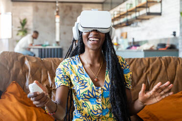 Positive African American female in VR goggles smiling and interacting with virtual reality while sitting on sofa over blurred cafe background in daylight - ADSF44867