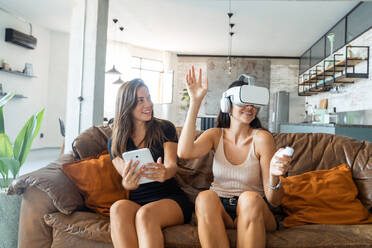 Cheerful young women with tablet phone and in VR goggles sitting on sofa while playfully exploring virtual reality cyberspace in living room - ADSF44868