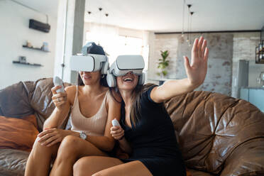 Cheerful young ladies in casual clothes with VR goggles sitting on sofa and interacting with virtual reality while entertaining at home - ADSF44870