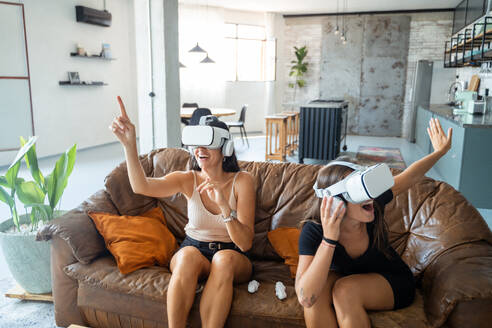 Cheerful young ladies in casual clothes with VR goggles sitting on sofa and interacting with virtual reality while entertaining at home - ADSF44871