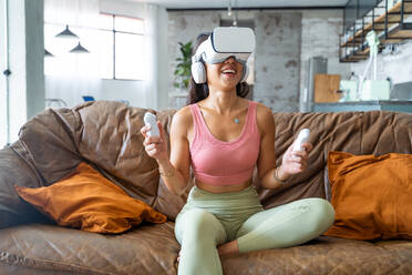 Young ethnic Asian female in VR goggles and with remote controllers playing video game while sitting on sofa in living room - ADSF44873