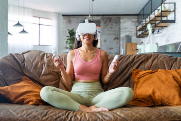 Young ethnic Asian female in VR goggles and with remote controllers playing video game while sitting on sofa in living room - ADSF44874