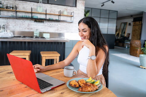 Positive young attractive woman in casual clothes sitting and browsing laptop at kitchen table with cup of drink eating healthy plate of toasts and croissant - ADSF44875