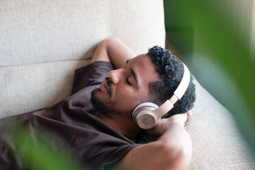 From above relaxed ethnic African American male in casual t shirt lying on comfortable sofa with closed eyes while listening to music on wireless headphones during rest at home - ADSF44881