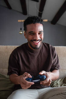 Happy young ethnic African American male in casual wear smiling and playing video game with joystick while chilling on comfortable bed at home during weekend - ADSF44883