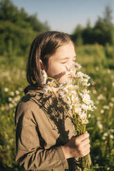 Junge mit geschlossenen Augen riecht an einem Strauß Gänseblümchen im Feld - ANAF01729