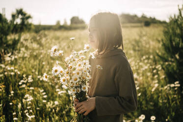 Contemplative boy holding bunch of daisies in field - ANAF01730