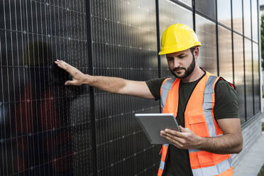 Engineer wearing hardhat examining solar panels through tablet PC - UUF29348