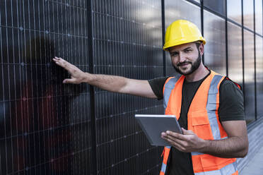 Smiling engineer holding tablet PC standing near solar panels - UUF29349