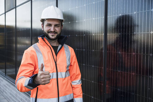 Lächelnder Ingenieur mit Daumen-hoch-Geste in der Nähe von Solarzellen - UUF29357