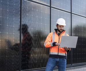 Engineer working on laptop standing near solar panels - UUF29362