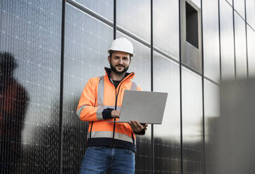 Smiling engineer holding laptop standing near solar panels - UUF29366