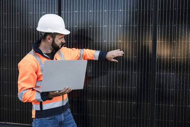Engineer wearing hardhat examining solar panels holding laptop - UUF29368