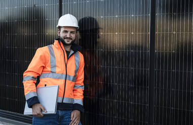 Smiling engineer leaning on solar panels holding laptop - UUF29371