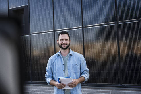 Smiling engineer holding notebook standing in front of solar panels - UUF29372