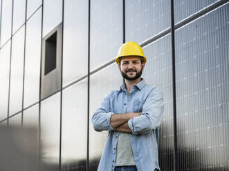 Lächelnder Ingenieur mit Schutzhelm, der mit verschränkten Armen vor einem Solarpanel steht - UUF29375