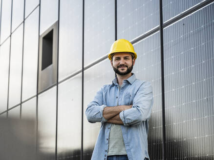 Smiling engineer wearing hardhat standing with arms crossed in front of solar panels - UUF29375